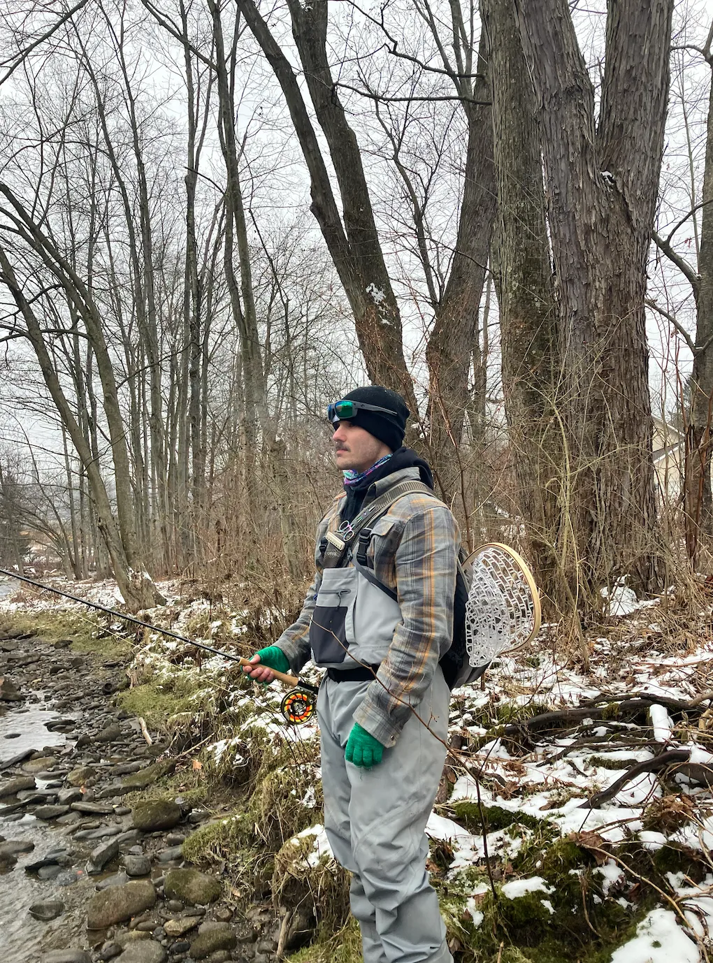Andrew standing at the edge of a snow-lined stream in January, rod in hand and a wooden net on his back.